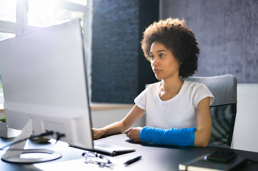 A young African-American woman sits at a computer. Her left arm is broken. She is looking for assistance in filing a claim.
